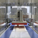 Zbliżenie Automatycznej pakowarki flow-pack SR550LR zgrzewającej plastikowe torebki na przenośniku taśmowym, z widocznymi w tle częściami mechanicznymi i metalowymi komponentami.