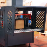 Kartonowe pudełko znajduje się wewnątrz Automatycznej formierki Speed Former 50 SB TN HM, dużej przemysłowej maszyny pakującej z czarną metalową ramą i ukośnymi bocznymi otworami wentylacyjnymi, znajdującej się na podłodze fabryki.