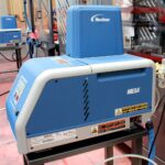 Niebiesko-szary dozownik kleju termotopliwego Nordson MESA na stojaku, z etykietami ostrzegawczymi i złączami węży, pokazany na targach - kompatybilny z Automatyczną zaklejarką Matic Tape 50 CF HM.