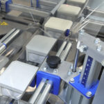 Zbliżenie na Automat pakujący poziomy FLOWPACK SI750S owijający prostokątne białe pojemniki przezroczystą folią plastikową na przenośniku, z widocznymi metalowymi i niebieskimi elementami.