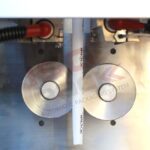 Zbliżenie na Pionową maszynę pakującą Mini Vert 240 PP, z dwoma metalowymi rolkami dociskającymi białą rurkę z nadrukowanym tekstem. W górnych rogach widoczne są czerwone i czarne przewody.