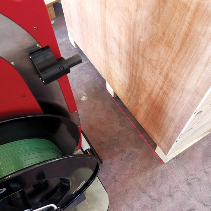 Zielona rolka taśmy i drewniana skrzynia leżą na podłodze. Pomiędzy nimi biegnie czerwona laserowa prowadnica, prawdopodobnie do wyrównania z Automatyczną taśmownicą do palet Flexy Strap T550.