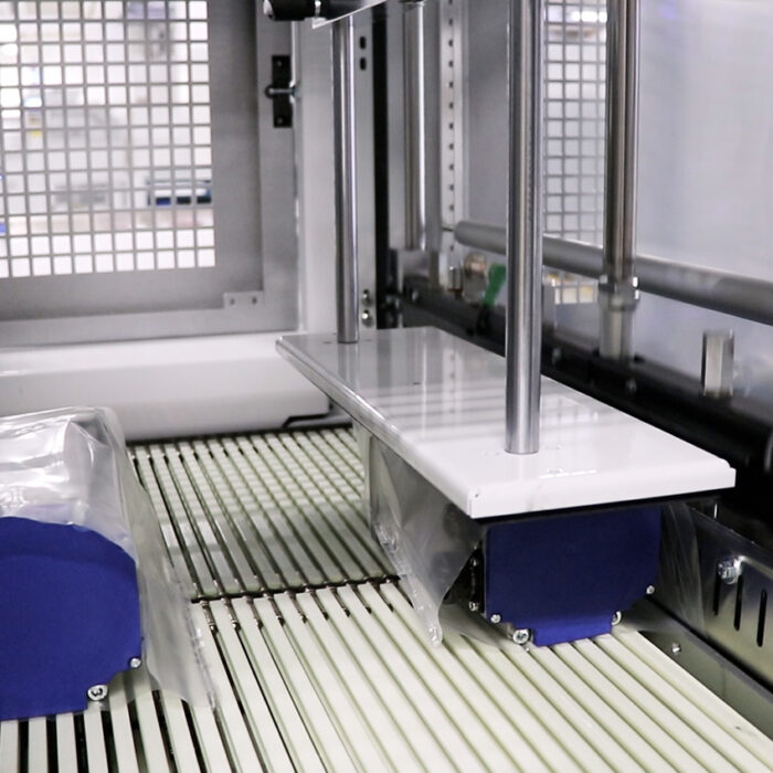 Zbliżenie na Automatyczną zgrzewarkę do zgrzewek FA802AS z przenośnikami taśmowymi przenoszącymi przezroczyste przedmioty owinięte plastikiem, metalowymi prętami i kratowanym ekranem bezpieczeństwa widocznym w tle.