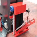 Zbliżenie czerwono-czarnego komponentu z palety Automatyczna owijarka Ultra Wrap 2000 BWF pokazuje niebieską wentylowaną pokrywę silnika, metalowe pręty i przewody na czerwonawej podłodze obok innych maszyn.