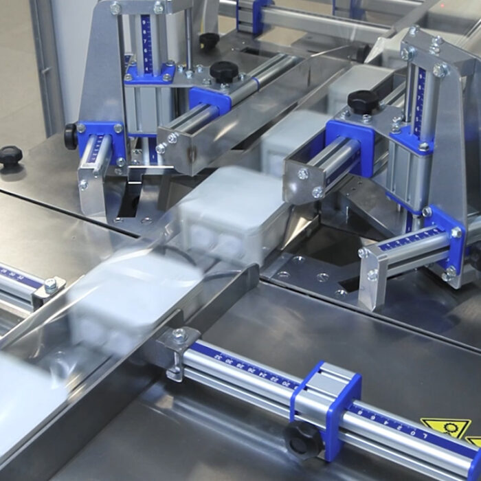 Zbliżenie na Automat pakujący poziomy FLOWPACK SBM750S, wyposażony w metalowe i niebieskie części, owijający prostokątne produkty w przezroczystą folię z tworzywa sztucznego na linii produkcyjnej.