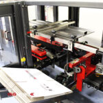 Zbliżenie Automatycznej formierki Speed Former 50 SB Servo pokazuje czerwone i metalowe części, stos płaskich kartonów gotowych do przetworzenia oraz logo LGS w dolnym rogu.