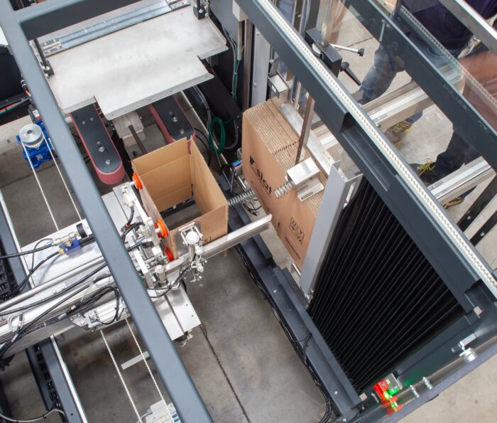 Widok z góry Automatycznej formiarki kartonów SIAT EZ-BOX XL pokazuje jej mechaniczne ramiona umieszczające płaski karton w otwartym pojemniku na hali fabrycznej, otoczonym przenośnikami taśmowymi i metalowymi ramami.
