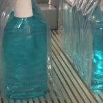 Zbliżenie kilku dużych butelek z niebieskim płynem, każda owinięta przezroczystym plastikiem przez Automatyczną zgrzewarkę do zgrzewek FA800AR-150Z i ustawiona na przenośniku taśmowym w otoczeniu przemysłowym.