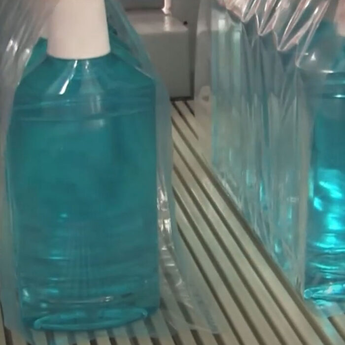 Zbliżenie kilku dużych butelek z niebieskim płynem, każda owinięta przezroczystym plastikiem przez Automatyczną zgrzewarkę do zgrzewek FA800AR-150Z i ustawiona na przenośniku taśmowym w otoczeniu przemysłowym.