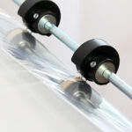 Zbliżenie dwóch metalowych prętów z czarnymi plastikowymi rolkami przytrzymującymi i prowadzącymi przezroczystą plastikową folię pod kątem, jak widać w Zgrzewarce kloszowej do folii Easy Sealer 85 podczas rozwijania folii.