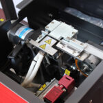 Zbliżenie maszyny przemysłowej podkreślającej Półautomatyczną bandownicę stołową Eco Strap 501, z widocznymi etykietami ostrzegawczymi, przewodami, przekładniami, silnikiem i naklejkami bezpieczeństwa wskazującymi ostrożność i ruchome części.