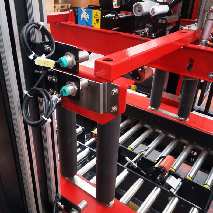 Zbliżenie systemu przenośników z czarnymi rolkami, czerwoną metalową ramą i srebrnymi czujnikami, zintegrowanego z Automatyczną zaklejarką Matic Tape 50 CF do automatycznego pakowania lub przenoszenia materiałów w fabryce.