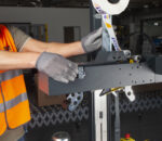 Pracownik w kamizelce ochronnej i szarych rękawicach reguluje półautomatyczną zaklejarkę kartonów NASTRO-SB - R5-R6, ładując zadrukowane etykiety na rolki w środowisku przemysłowym.