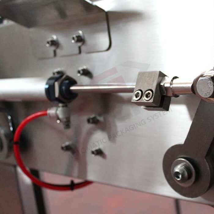 Zbliżenie na zespół mechaniczny ze stali nierdzewnej ze śrubami, wałem i czerwonym kablem na tle szczotkowanego metalu. To urządzenie jest częścią Półautomatycznej formierki Easy Form 50 SS do zastosowań przemysłowych i pakowania.