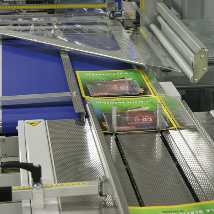 Dwa pudełka DVD przesuwają się na metalowym przenośniku w Automatycznej zgrzewarce kątowej Ultra Sealer 80 HSS, z plastikową owijką częściowo zakrywającą jedno pudełko. Scena rozgrywa się w zautomatyzowanym zakładzie pakowania.