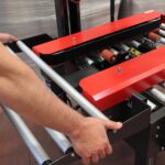 Osoba reguluje metalową ramę i czerwono-czarne rolki Półautomatycznej zaklejarki Eco Tape 50 SB, przygotowując ją do pracy w środowisku produkcyjnym lub wysyłkowym.