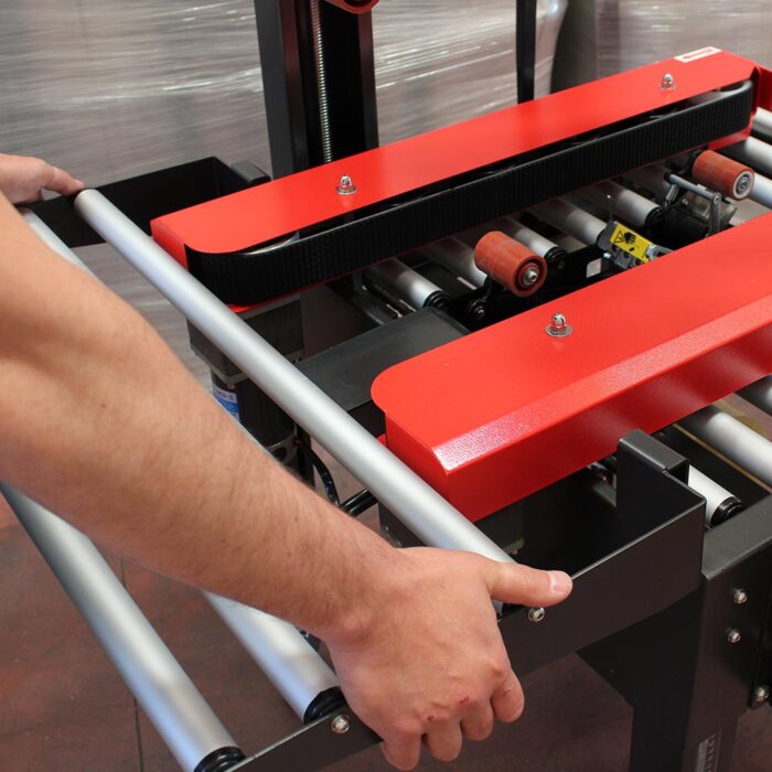 Osoba reguluje metalową ramę i czerwono-czarne rolki Półautomatycznej zaklejarki Eco Tape 50 SB, przygotowując ją do pracy w środowisku produkcyjnym lub wysyłkowym.