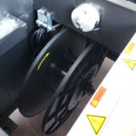 Zbliżenie czarnej szpuli na Półautomatyczna bandownica stołowa Eco Strap 501 C, z żółtą strzałką kierunkową, widocznymi częściami maszyny i etykietami ostrzegawczymi z symbolami ostrzeżenia i niebezpieczeństwa.