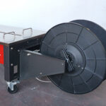 Zbliżenie na dużą czarną plastikową szpulę na metalowej ramie na kółkach, przymocowaną do paska Półautomatyczna bandownica stołowa Eco Strap 501L w kolorze czerwonym i srebrnym, ustawiona na betonowej podłodze przy gładkiej ścianie.