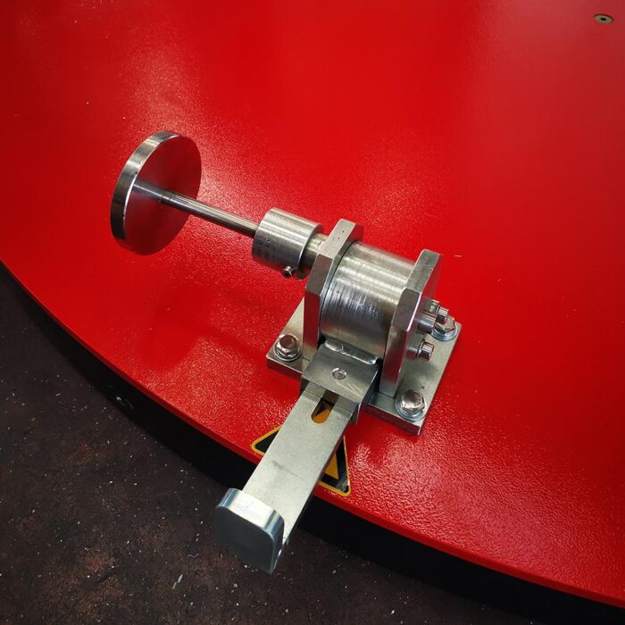 Zbliżenie metalowego elementu z palety Półautomatyczna owijarka Easy Wrap 2000 B RC, zamontowanego na jasnoczerwonej powierzchni, z cylindrycznym korpusem, płaskim dyskiem i długą prostokątną dźwignią zabezpieczoną śrubami.
