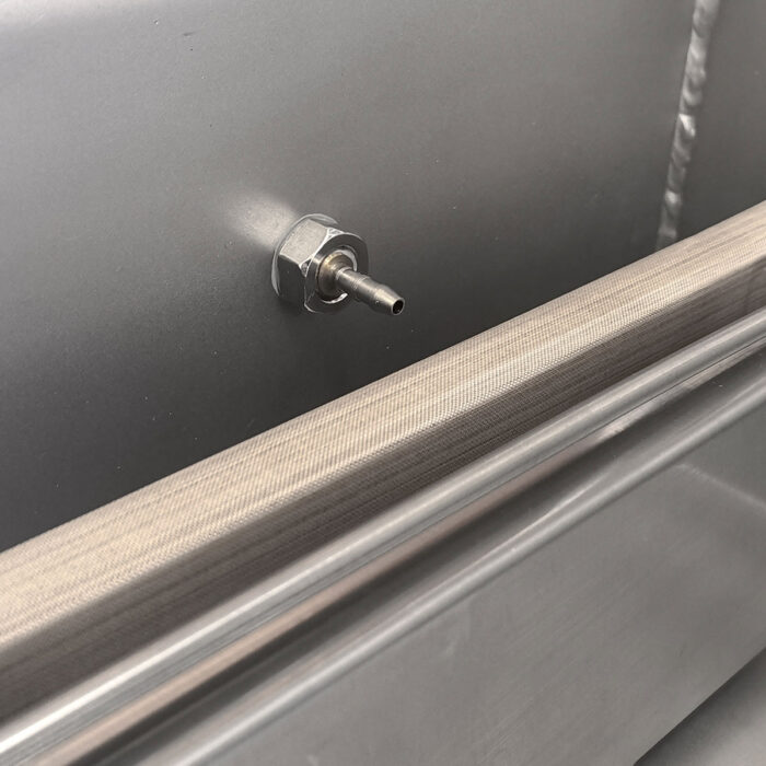 Zbliżenie metalowej armatury z dyszą na szarej powierzchni przemysłowej obok taśmy siatkowej i metalowych paneli - elementów Komorowej pakowarki próżniowej Eco Vac 300S TB.