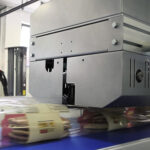 Zbliżenie na Automat pakujący poziomy FLOWPACK SBM750S-LR zgrzewający zapakowane w folię paczki ciastek, gdy poruszają się one wzdłuż niebieskiego przenośnika taśmowego w fabryce.