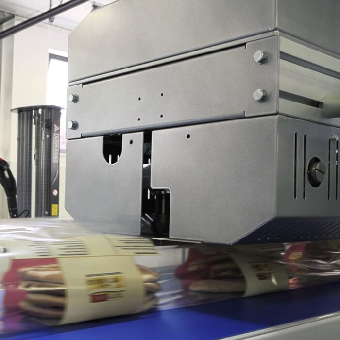 Zbliżenie na Automat pakujący poziomy FLOWPACK SBM550S-LR zgrzewający przezroczysty plastik wokół paczek ciastek na przenośniku taśmowym w fabryce.