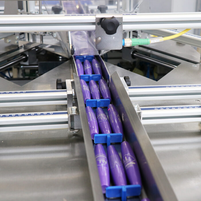 Zbliżenie na Automat pakujący poziomy FLOWPACK SI550S-LR transportujący kilka fioletowych cylindrycznych produktów w rzędzie po metalowej szynie w warunkach fabrycznych.