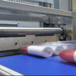 Zbliżenie na Automatyczną pakowarkę flow-pack SR750LR w fabryce, z niebieskim przenośnikiem szybko przesuwającym czerwone i białe cylindryczne elementy obok metalowej jednostki tnącej lub zgrzewającej.