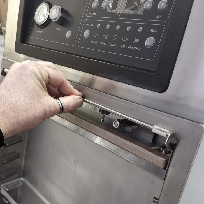 Dłoń nosząca srebrną obrączkę pociąga za metalową dźwignię na panelu Komorowej pakowarki próżniowej Eco Vac 420V TB, wyposażonym w przyciski, wskaźniki i ekran wyświetlacza.