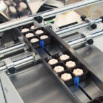 Plastikowe tacki z ciastkami w kształcie kwiatów przesuwają się wzdłuż linii produkcyjnej wyposażonej w maszyny metalowe i Automatyczną pakowarkę flow-pack SR750 w zakładzie przetwórstwa spożywczego.