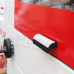 Zbliżenie czarno-białego uchwytu na czerwono-białej automatycznej zgrzewarce kątowej do folii Matic Sealer 65 E, z czarnym okrągłym pokrętłem po lewej stronie. Napis AUTOMATIC PACKAGING SYSTEM jest niewyraźny.