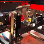 Zbliżenie na maszynę Automatyczna formierka Speed Former 50 SB TN HM z metalowymi elementami, śrubami, pomarańczowymi kablami, manometrem i etykietą ostrzegawczą na czerwono-czarnej ramie w warsztacie.