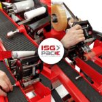 Zbliżenie rąk regulujących rolki, pasy i dyspenser taśmy na automatycznej maszynie pakującej Półautomatyczna zaklejarka Random Tape 50 TBB w kolorze czerwonym, z widocznym logo ISG PACK podczas pracy maszyny.