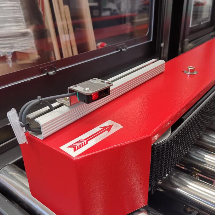 Zbliżenie czerwonej części przemysłowej na Półautomatyczną zaklejarkę Random Tape 50 SB, pokazujące zamontowany czujnik, naklejkę ze strzałką skierowaną w prawo i widoczne rolki z innymi elementami maszyny w tle.