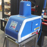 Niebieska Automatyczna zaklejarka Auto Tape 50 HM z cyfrowym panelem sterowania stoi na metalowym stojaku w fabryce, otoczona różnymi urządzeniami.