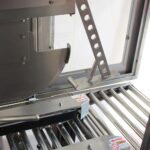 Zbliżenie na Automatyczną zaklejarkę Auto Tape 50 CF SS, maszynę przemysłową ze stali nierdzewnej z rolkami i etykietami bezpieczeństwa, używaną do przetwarzania lub przenoszenia materiałów w produkcji lub produkcji żywności.