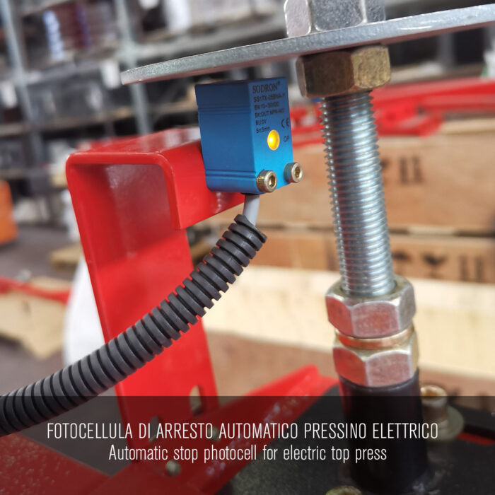 Zbliżenie niebieskiej fotokomórki automatycznego zatrzymania zamontowanej na metalu z czerwonym wspornikiem, do użytku z Półautomatyczną owijarką paletową PRE-STRETCH Easy Wrap 2000 BC. Nakładka tekstowa w języku włoskim i angielskim. Tło przemysłowe.