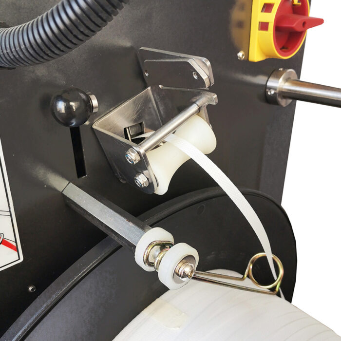 Zbliżenie na Automatyczną taśmownicę do palet Easy Strap 309/312, pokazującą białą taśmę PP przechodzącą przez rolki i prowadnice, z metalowymi i plastikowymi częściami oraz przyciskami sterującymi na czarnej powierzchni maszyny.