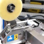 Zbliżenie na Półautomatyczną zaklejarkę Eco Tape 50 SB SS z dużą rolką przezroczystej taśmy i metalowymi częściami, pokazujące etykiety ostrzegawcze i instruktażowe na korpusie ze stali nierdzewnej.