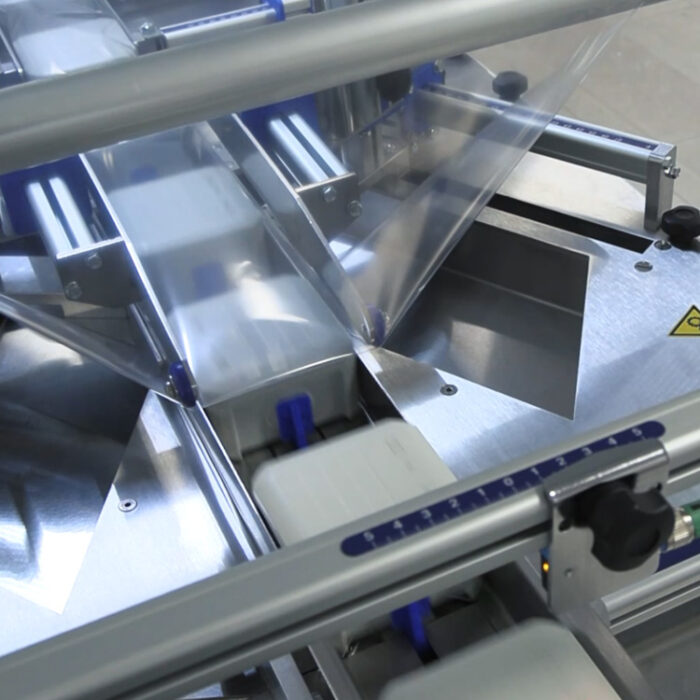 Zbliżenie na Automat pakujący poziomy FLOWPACK SBM550S owijający prostokątne produkty w folię plastikową na przenośniku taśmowym, z metalowymi i niebieskimi elementami oraz widocznym znakiem ostrzegawczym.
