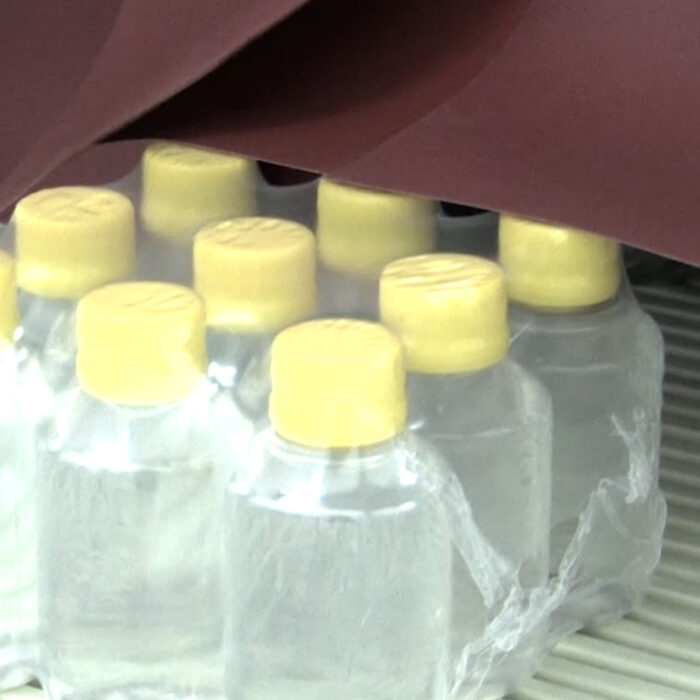 Przezroczyste plastikowe butelki z żółtymi nakrętkami są pakowane w przezroczystą folię termokurczliwą przez Automatyczną zgrzewarkę do zgrzewek FA600AR-150R i umieszczane na przenośniku taśmowym.