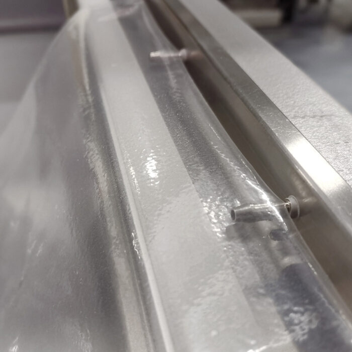 Zbliżenie przezroczystego plastikowego arkusza zgrzanego z metalem z wystającymi małymi dyszami, prawdopodobnie część procesu przemysłowego z użyciem Komorowej pakowarki próżniowej Ultra Vac 600 SA.