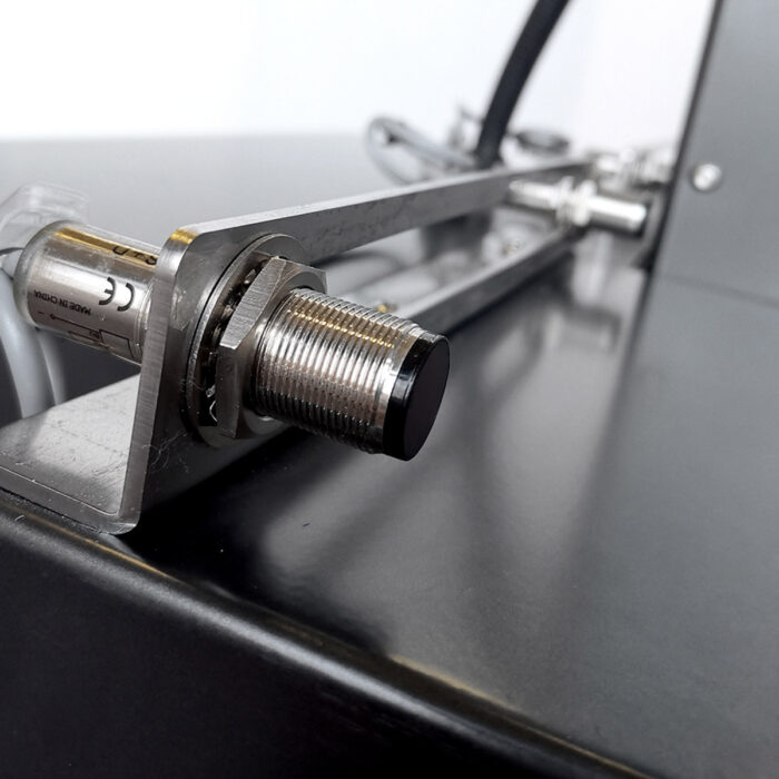 Zbliżenie metalowego cylindrycznego czujnika lub przełącznika zamontowanego na wsporniku przymocowanym do sprzętu przemysłowego, takiego jak Bandownica ramowa wiązarka PP Easy Strap 309BP/312BP, z przewodami połączeniowymi widocznymi w tle.