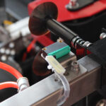 Zbliżenie maszyny przemysłowej przedstawia metalowy czujnik z zielonymi i czarnymi elementami zamontowany na Automatycznej Formierce Prędkości 50 SB TN HM, z czerwonymi kablami i okrągłym ciemnym dyskiem w tle.