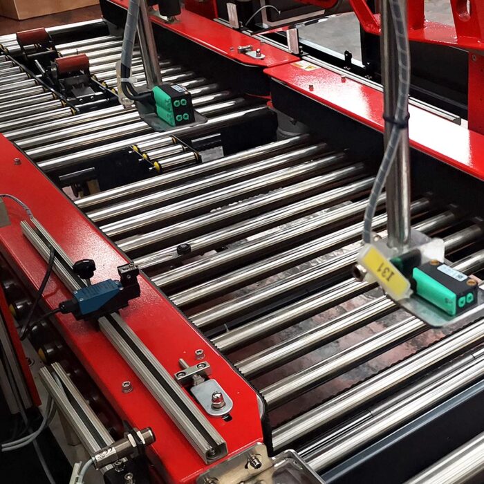 Zbliżenie metalowego przenośnika rolkowego z czerwoną ramą i czujnikami, idealnego do automatyzacji ruchu przedmiotów i śledzenia za pomocą Automatycznej zaklejarki Matic Tape 50 CF w fabryce.