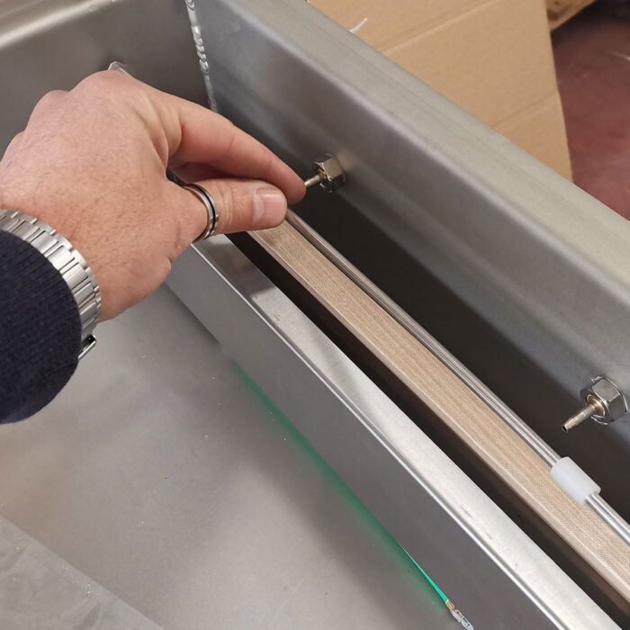 Osoba reguluje metalową śrubę w Komorowej pakowarce próżniowej Eco Vac 300S TB, trzymając pasek drewna między dwiema metalowymi częściami, mając na sobie srebrny zegarek i pierścionek.