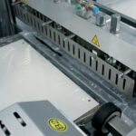 Zbliżenie na Automatyczną zgrzewarkę kątową Ultra Sealer 80 SM w użyciu, zgrzewającą plastikowy arkusz pod podgrzewaną listwą. Na metalowych elementach widoczne są etykiety ostrzegające przed gorącymi powierzchniami.