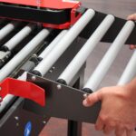 Zbliżenie przedstawia ręce regulujące szary przenośnik rolkowy z metalowymi rolkami i czerwoną częścią, używany do przenoszenia paczek w warunkach przemysłowych za pomocą półautomatycznej zaklejarki Eco Tape 50 SB.