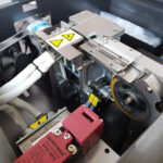 Zbliżenie na Półautomatyczną bandownicę stołową Eco Strap 501L, ukazujące metalowe koła zębate, etykiety ostrzegawcze, wentylator chłodzący, wyłączniki bezpieczeństwa, kable i ruchome części mechaniczne wewnątrz obudowy.